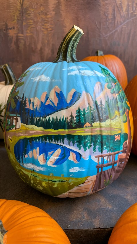 A pumpkin with a painting of a serene landscape. The landscape features a calm lake reflecting the surrounding mountains, trees, and a wooden deck.